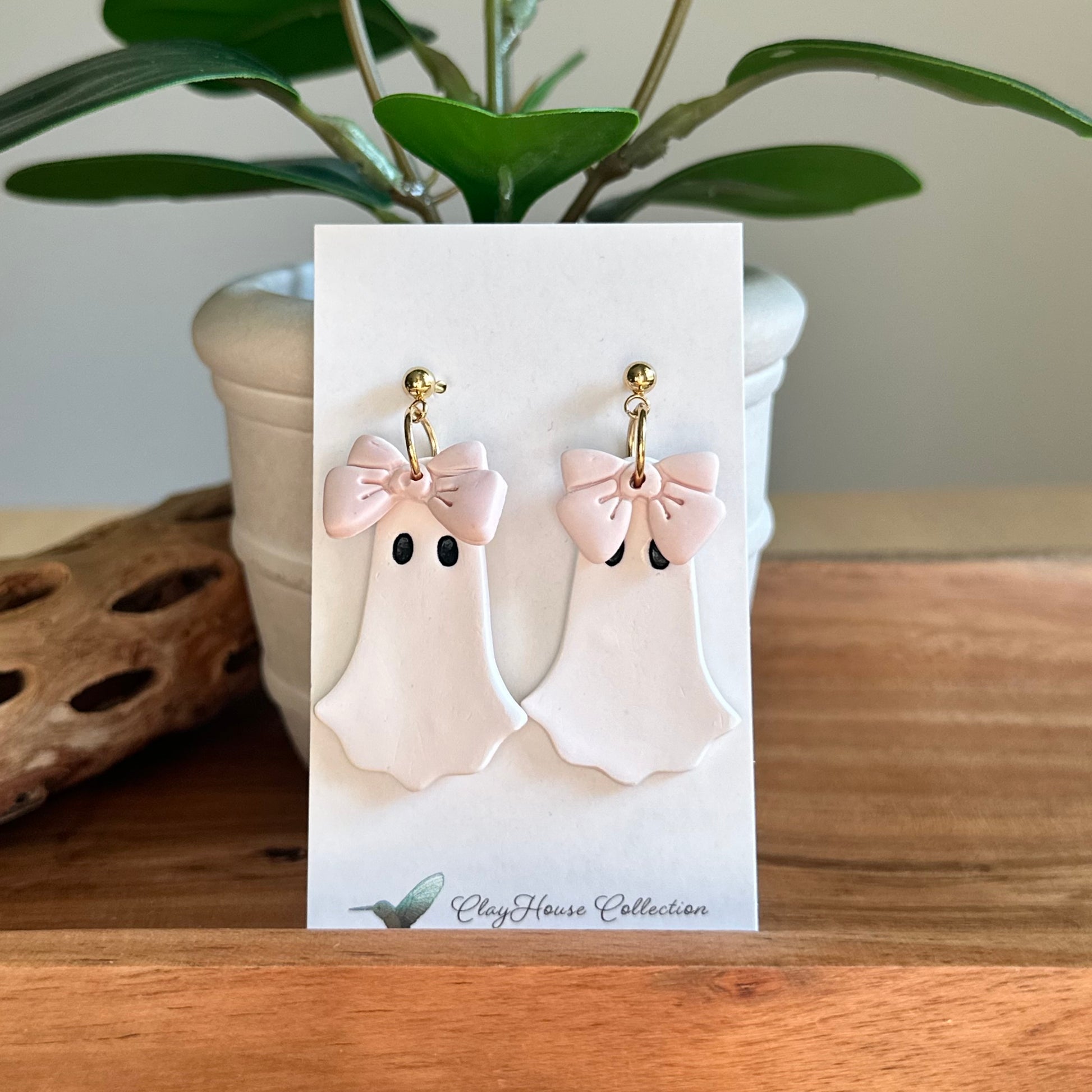 Ghost-shaped earrings with pink bows on a card, placed on a wooden surface with a plant in the background.