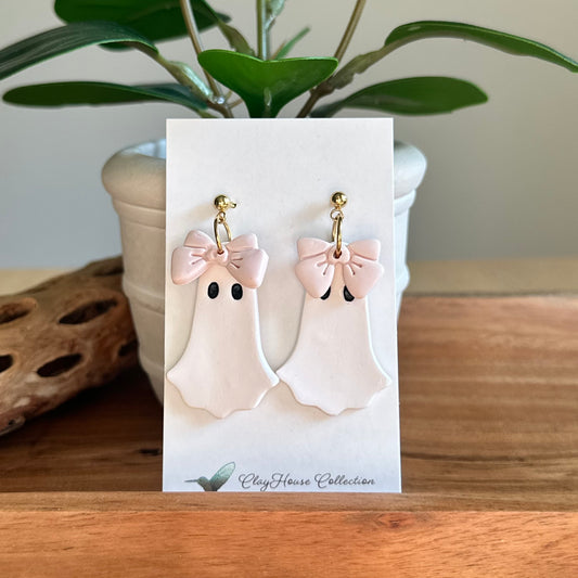 Ghost-shaped earrings with pink bows on a card, placed on a wooden surface with a plant in the background.