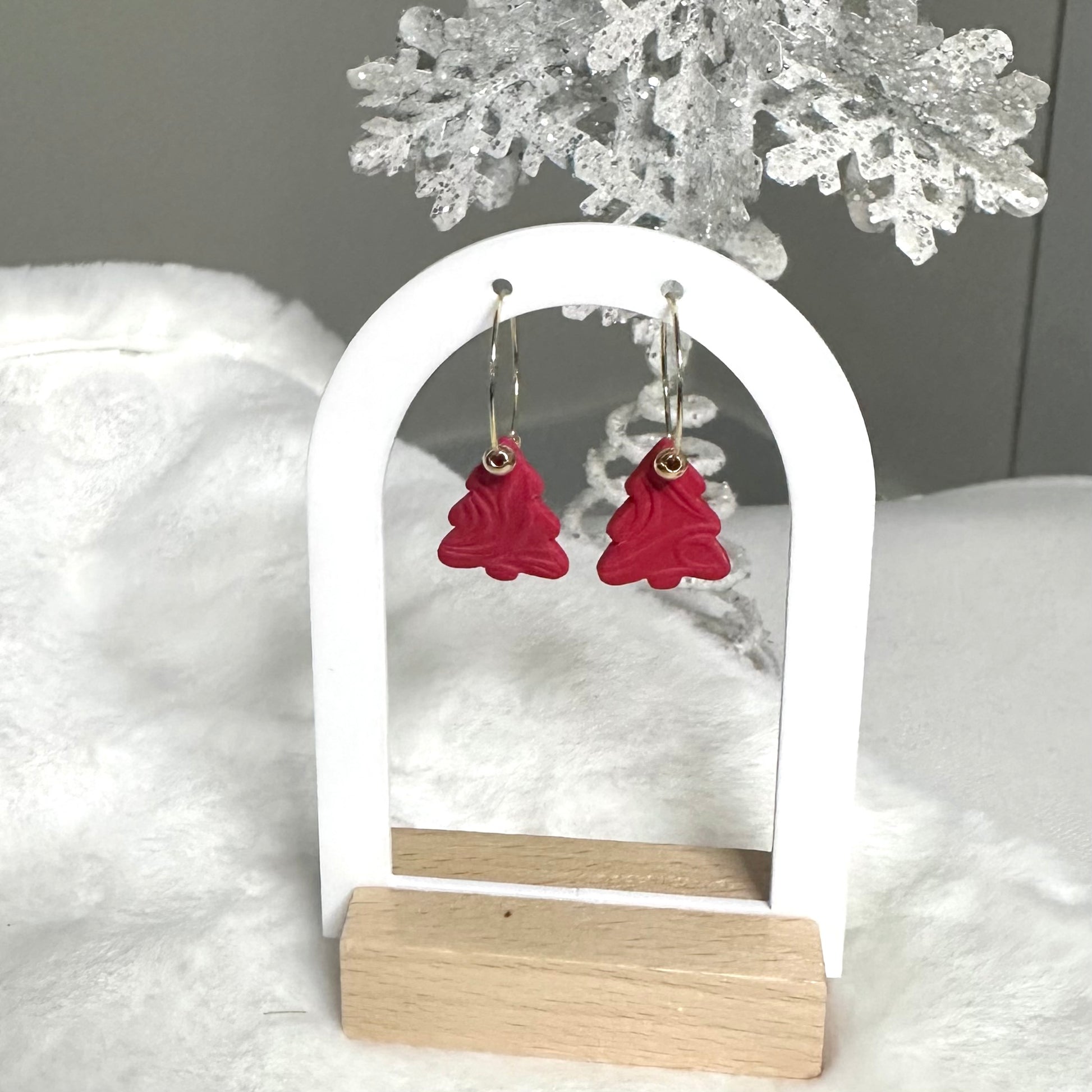 Red Christmas tree-shaped earrings on a white and wooden stand with a snowy background.