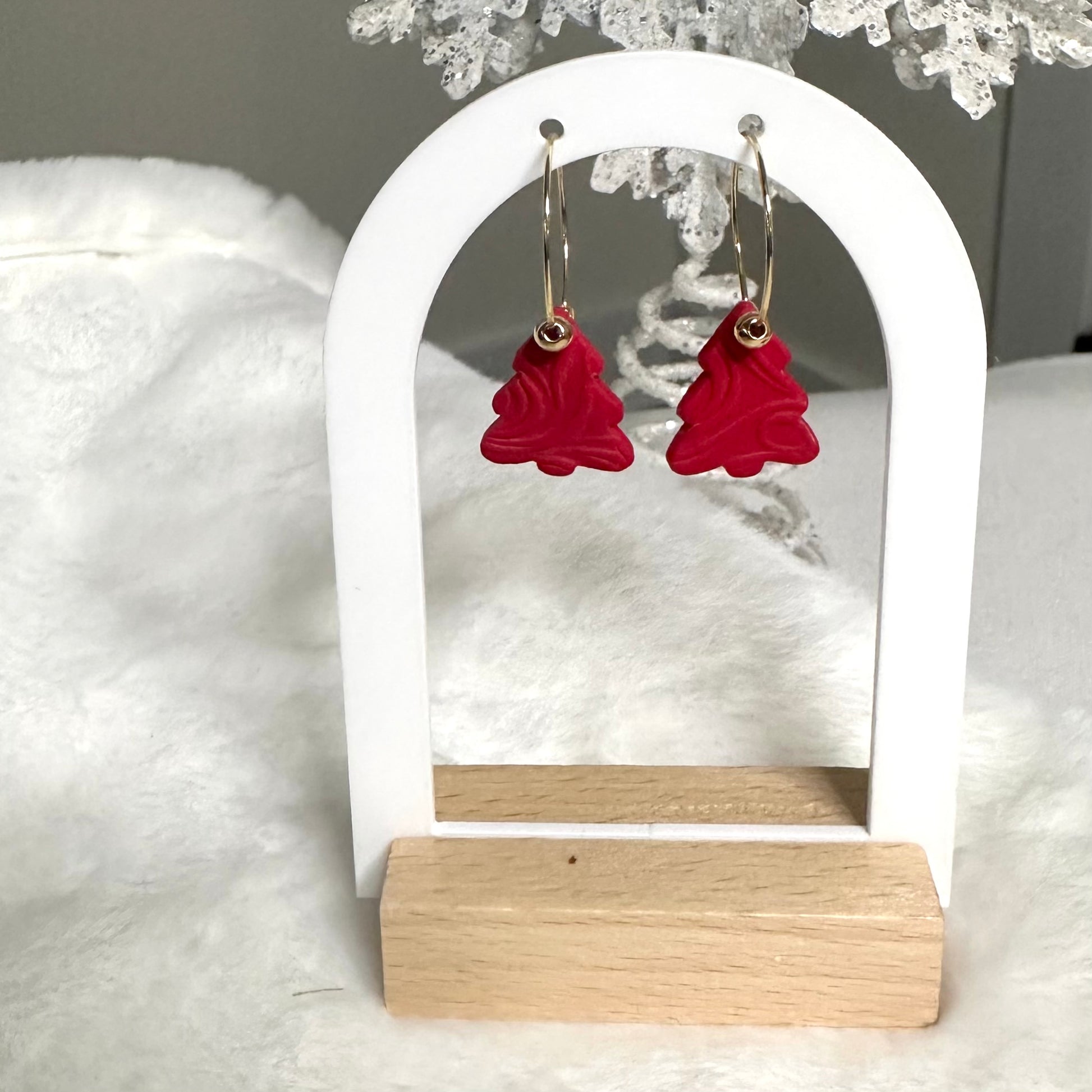 Red Christmas tree-shaped earrings on a white and wooden stand with a snowy background.