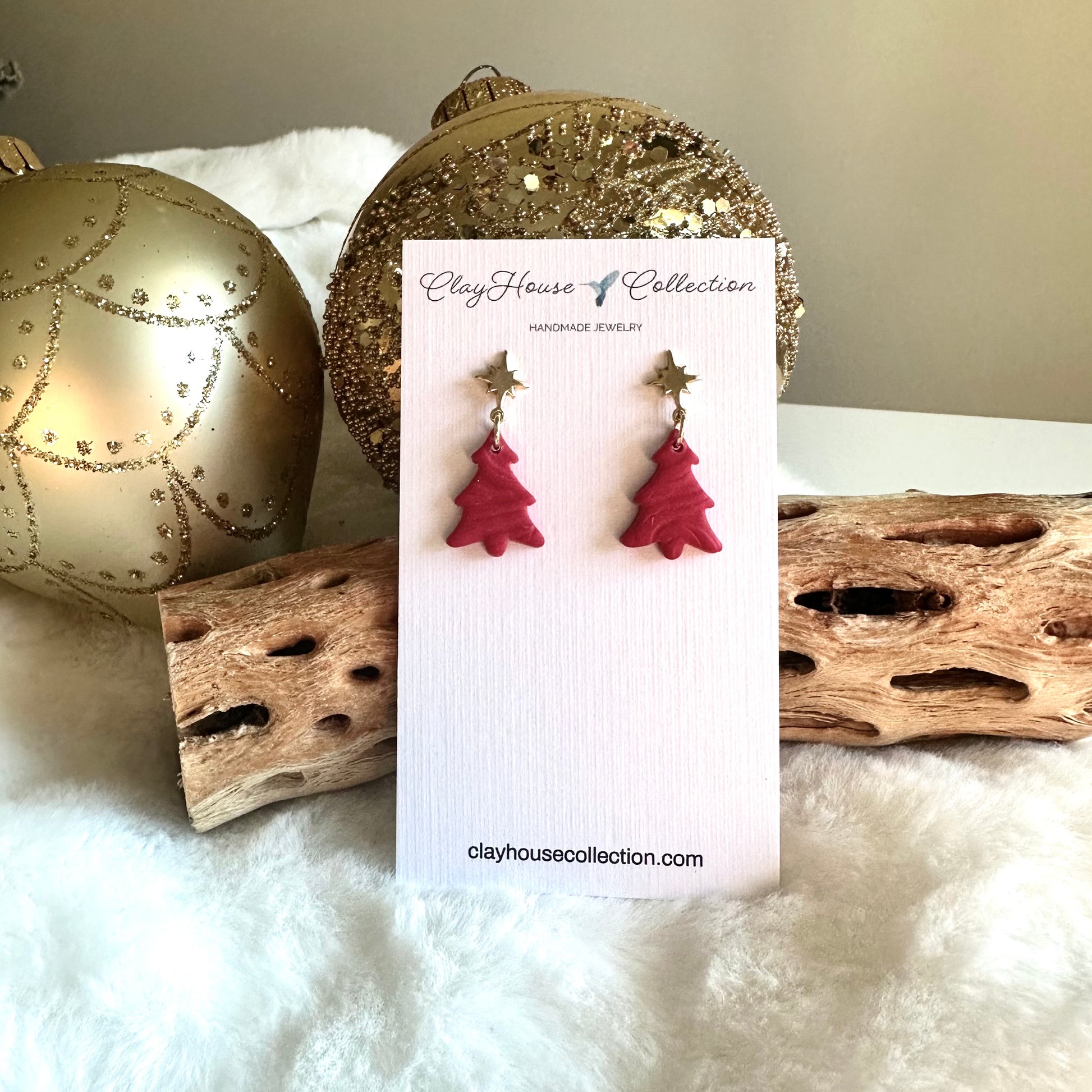 Red Christmas tree earrings on a Clay House Collection card with decorative items in the background.